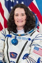 A white woman with long dark hair in an astronaut's spacesuit without the helmet is shown against an American flag.