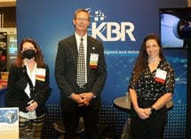Two women stand on either side of a man. Behind them is a large sign in dark blue with the logo for KBR, the exhibitor, in white.