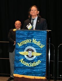A man in a black suit and pale blue shirt with a dark tie stands behind a podium with a mounted microphone. Another gentleman stands behind and to his right wearing a dark jacket and light pants. His hands are clasped in front of him. A clothdeep blue sign showing AsMA's logo in color and the Association's name in yellow is hung on the front of the podium.