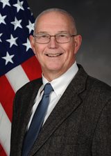 A white man with closely cropped pale hair wearing glasss, a white shirt, dark grey pinstripe jacket, and blue tie is shown against a dark grey backround with part of an American flag on the left.