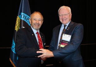 On the left is Gary Gray, who received his 50-year pin from AsMA President Jim DeVoll on the right.They are shaking hands while smiling at the camera.