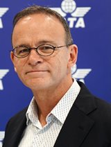 A white man is shown wearing glasses, a white and light grey pinstriped shirt, and black jacket against a medium blue background with IATA's logo.