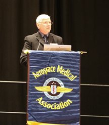 A white-haired gentleman with glasses stands behind a podium draped with a dark blue flag with AsMA's logo in color and its name in yellow. He is speaking into the attached microphone and there is a laptop on the podium in front of him.