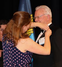 A copper-haired woman, seen from the back and wearing a dark blue dress with pink polka dots puts the Past President's pin on Dr. DeVoll, on the right, a white-haired man with glasses in a black suit, white shirt, and black bow tie.