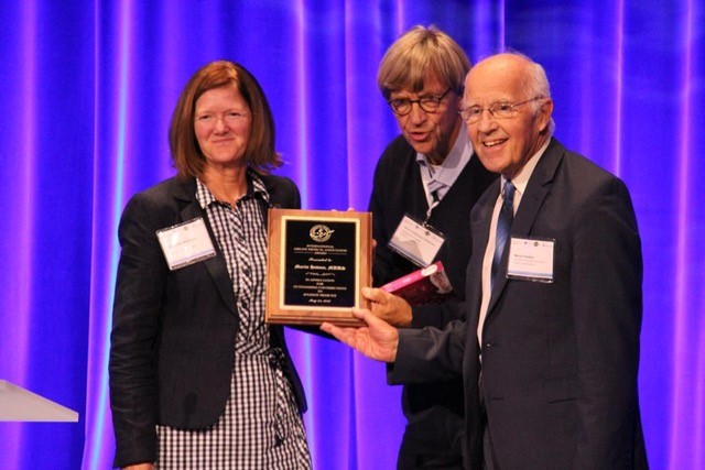 A white woman with coppery hair, left, stands with two white men with glasses. The middle one with blond hair and the one on the right partially bald with white hair. The gentleman on the right holds a wooden award plaque up that has a black inset with white lettering.