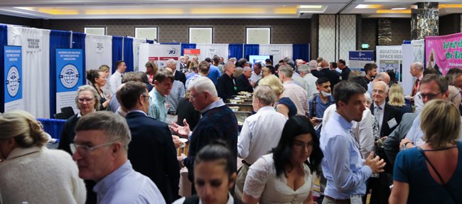 In a hotel ballroom with light colored walls, a group of people, both men and women, stand eating, drinking, and talking. Around them are blue and white curtains on frames with posters bearing the names of the exhibitors.