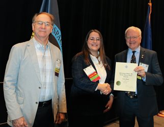 A woman, the Mohler scholarship winner, stands between AsMA President Jim DeVoll on her left and Kim Broadwell, representing the AsMA Foundation, on her right. Dr. DeVoll is holding up the certificate for the scholarship. The background is a black curtain and there are two flags behind the people.
