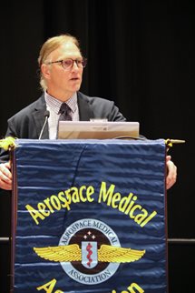 UHMS's President, a man, stands behind a podium with a mounted microphone. He is in a medium grey suit with a white shirt and dark tie. On the front of the podium is a deep blue cloth sign with AsMA's logo in color and its name in yellow.