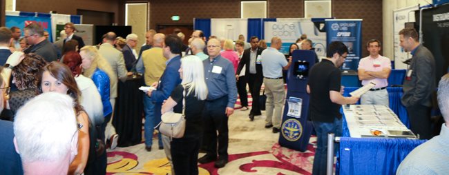 In a hotel ballroom with dark brown walls and light tan panels, people stand talking. Around them are blue and white curtains on frames with posters showing the names and information about the exhibitors. To the right are some tables with medium blue tablecloths spread with brochures and other items.