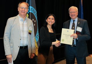 A dark-haired woman, the winner of the Mantri scholarship, stand between Kim Broadwell, representing the AsMA Foundation, on her right and Jim DeVoll, AsMA President, who is holding up the certificate, on her left. Behind them is a black curtain and two flags.