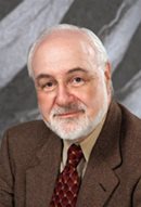 A white man with white hair and a white beard and mustache in a brown jacket, white shirt and polka dot maroon tie is shown against a grey and white marbled background.
